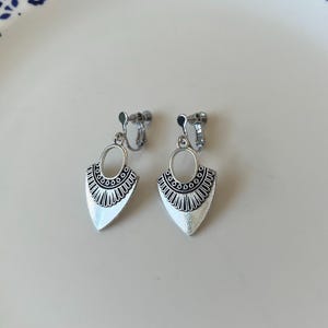 Può includere: Un paio di orecchini a clip argentati. Ogni orecchino presenta un pendente decorativo a forma di scudo con un ritaglio ovale e un design strutturato dettagliato. Gli orecchini sono esposti su una superficie bianca.