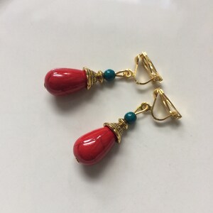 Könnte beinhalten: Ein Paar rote und goldene Ohrclips mit türkisfarbenen Perlen. Die Ohrringe haben einen tropfenförmigen roten Stein mit einem goldenen Akzent.