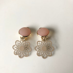 Pode incluir: Um par de brincos de pressão. Cada brinco apresenta um topo circular rosa claro com uma borda dourada. Abaixo, um design de flor vazada dourada pende. Os brincos são exibidos em uma superfície branca.