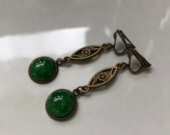 Puede incluir: Un par de pendientes de clip verdes y dorados con un diseño floral. Los pendientes presentan una piedra redonda verde con un acabado brillante.