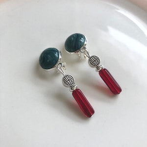 Könnte beinhalten: Ein Paar silberne Ohrclips mit einem dunkelblauen Wirbelmuster und einem roten Glasperlen-Anhänger.