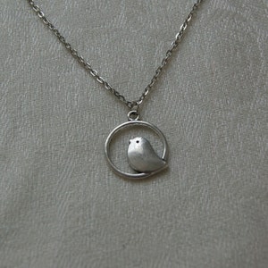 Pode incluir: Um colar de prata com um pequeno pingente de pássaro dentro de um círculo.