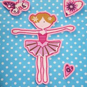 May include: A pink and brown ballerina applique with a tutu, arms outstretched, and two small flower accents in her hair. A butterfly and two heart-shaped appliques are also present, all on a blue polka dot background.