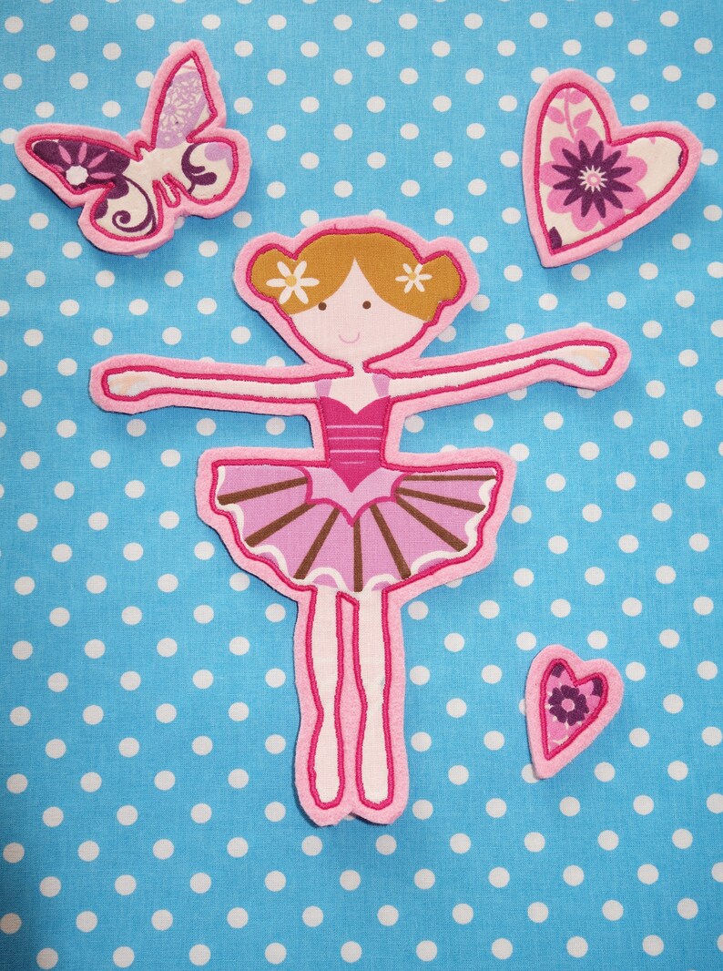 May include: A pink and brown ballerina applique with a tutu, arms outstretched, and two small flower accents in her hair. A butterfly and two heart-shaped appliques are also present, all on a blue polka dot background.