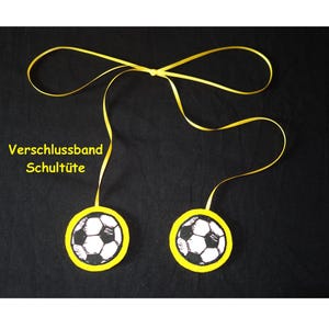 May include: Two circular decorations featuring a black and white football pattern with a bright yellow border, connected by a long yellow ribbon tied in a bow at the top. The text "Verschlussband Schultüte" is printed in yellow on the left side.