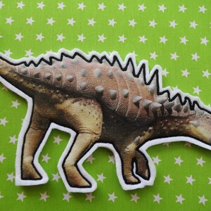 Puede incluir: Un parche de dinosaurio marrón y blanco con un contorno blanco. El dinosaurio tiene una fila de púas en la espalda y una cabeza grande con una boca ancha.