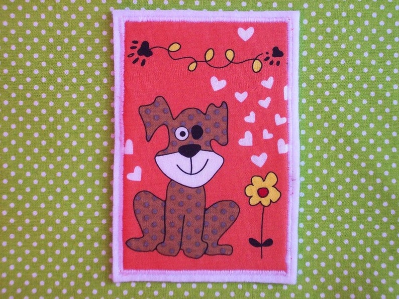 May include: A rectangular patch with a cartoon dog design. The dog is brown with spots, a white face, and a black nose, set against a red background. White hearts and a yellow flower are also present. The patch has a white border, on a green polka dot background.