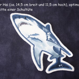 Könnte beinhalten: Eine weiße Filzapplikation eines blauen Hais mit weißem Umriss. Der Hai ist 14,5 cm breit und 11,5 cm hoch. Der Text "großer Hai (ca. 14,5 cm breit und 11,5 cm hoch), optimal für die Mitte einer Schultüte" ist über dem Hai sichtbar.