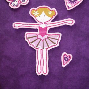 May include: Purple fabric featuring appliqued designs: a ballerina, a butterfly, and two hearts. The ballerina has a pink tutu and blonde hair. The butterfly and hearts have floral patterns.
