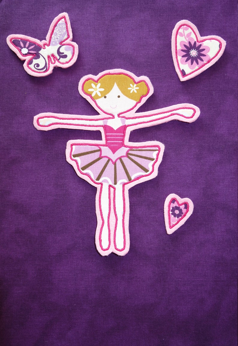 May include: Purple fabric featuring appliqued designs: a ballerina, a butterfly, and two hearts. The ballerina has a pink tutu and blonde hair. The butterfly and hearts have floral patterns.