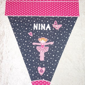 May include: A triangular pennant banner in pink and navy blue, decorated with white stars, a ballerina, and the name "NINA". The banner features a pink star pattern at the top and bottom, with a butterfly and hearts.
