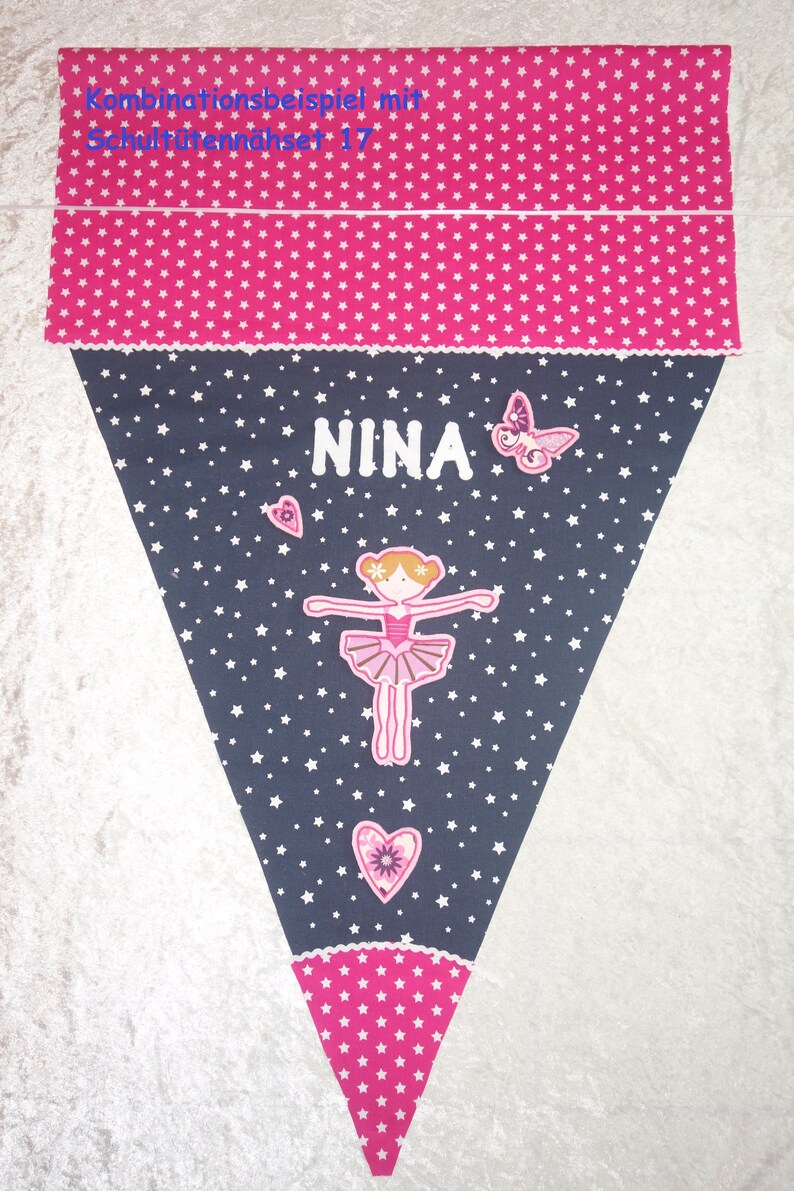 May include: A triangular pennant banner in pink and navy blue, decorated with white stars, a ballerina, and the name "NINA". The banner features a pink star pattern at the top and bottom, with a butterfly and hearts.