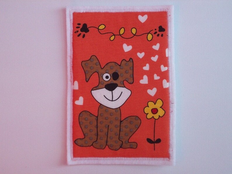 May include: An orange fabric patch featuring a cartoon dog illustration. The dog is brown with darker brown spots, a white face, and a black nose. White hearts and a yellow flower are also featured. The patch has a white frayed edge.