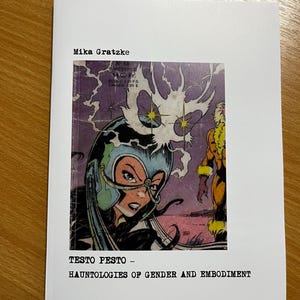 May include: A printed paper with a comic book illustration. The image features a woman in a helmet and a yellow-clad figure. The text includes "Mika Gratzke" and "TESTO PESTO - HAUNTOLOGIES OF GENDER AND EMBODIMENT."