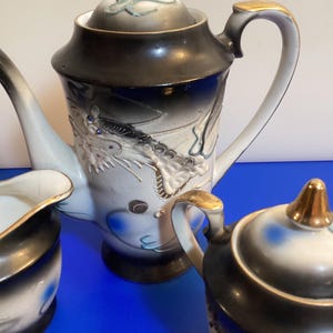 Op de afbeelding: Een porseleinen theeservies met een theepot, melkkan en suikerpot. De set is versierd met een drakenmotief in wit, zwart en blauw, met gouden accenten op de handvatten en deksels. De theepot heeft een lange, gebogen tuit.