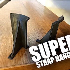May include: Two black 3D printed plastic strap hangers. The hangers are designed to hold straps vertically on a wall. The text "SUPER STRAP HANGER!" is superimposed on the image.
