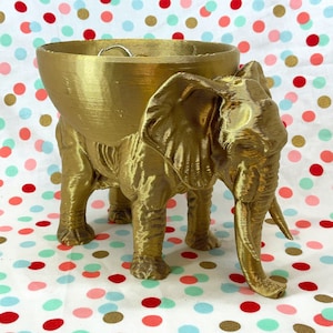 Puede incluir: Un macetero de elefante dorado con un tazón en la parte superior. El macetero está hecho de un material texturizado y tiene un diseño de elefante realista.