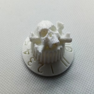 Op de afbeelding: Witte kunststofknop met een schedel en kruisbeenderen ontwerp. De knop heeft de nummers 1 tot en met 6 erop gedrukt.