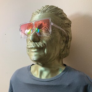 May include: A gold-colored bust of a man wearing clear framed sunglasses with pink lenses. The bust is wearing a dark grey t-shirt with a red circle graphic on the front.