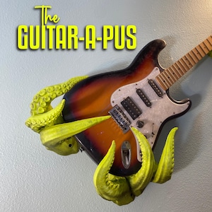 Op de afbeelding: Een gitaar met een felgele octopusvormige wandbevestiging. De tekst "The Guitar-A-Pus" staat in gele letters boven de gitaar.
