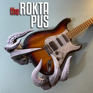 以下が含まれることがあります： 白ピックガード付きの茶色と白のエレキギターが、灰色のタコ型のギタースタンドを使って壁に取り付けられています。画像の左上隅に「the ROKTA PUS」というテキストが表示されています。
