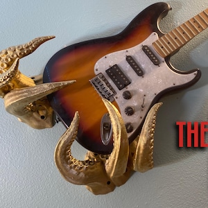 May include: A sunburst electric guitar with a white pickguard is mounted on a wall using a decorative golden octopus-like sculpture. The text "THE ROKTA PUS" is visible in the lower right corner of the image.