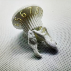 Peut inclure: Tour de dés en plastique blanc imprimé en 3D avec un design de crâne et les chiffres 4, 5, 6, 7, 8 et 9 imprimés sur le dessus.