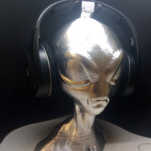May include: Black headphones with a silver alien head stand. The headphones are on the stand, and the stand is on a white surface.