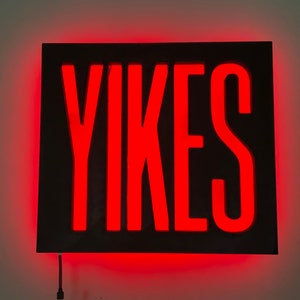 Puede incluir: Un letrero rectangular negro con la palabra "YIKES" en letras de neón rojas.
