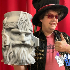 May include: A gray 3D-printed bust of a bearded figure with a helmet. The bust is held in front of a red curtain. The bust has intricate details and a stern expression. The bust is a decorative item.