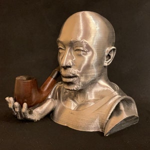 May include: A silver bust of a person with a pipe in their hand. The bust is made of a 3D printed material and has a smooth, metallic finish.