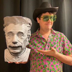 May include: A person wearing a black cowboy hat and green sunglasses is holding a white 3D printed bust of Albert Einstein.