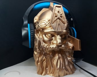 Könnte beinhalten: Ein goldfarbener 3D-gedruckter Kopfhörerständer in Form eines bärtigen Kriegerkopfes. Der Ständer hat ein blaues und schwarzes Headset darauf.