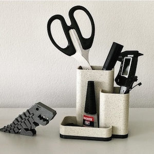 May include: A white desk organizer with a speckled finish holds a pair of black scissors, a black pen, a black measuring tool, and a black bottle of glue. A gray dinosaur figurine sits on a white surface in front of the organizer.