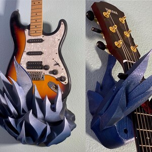 Puede incluir: Un soporte de pared para guitarra impreso en 3D de color azul con un diseño de púas. El soporte sostiene una guitarra con una pala negra y clavijas de afinación doradas. El texto "YAMAHA" es visible en la pala.