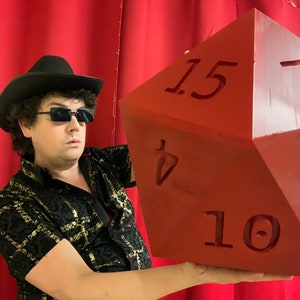 Puede incluir: Una persona con un sombrero negro y gafas de sol sostiene un gran dado rojo de 20 caras con los n&uacute;meros 10, 15 y 7 visibles.