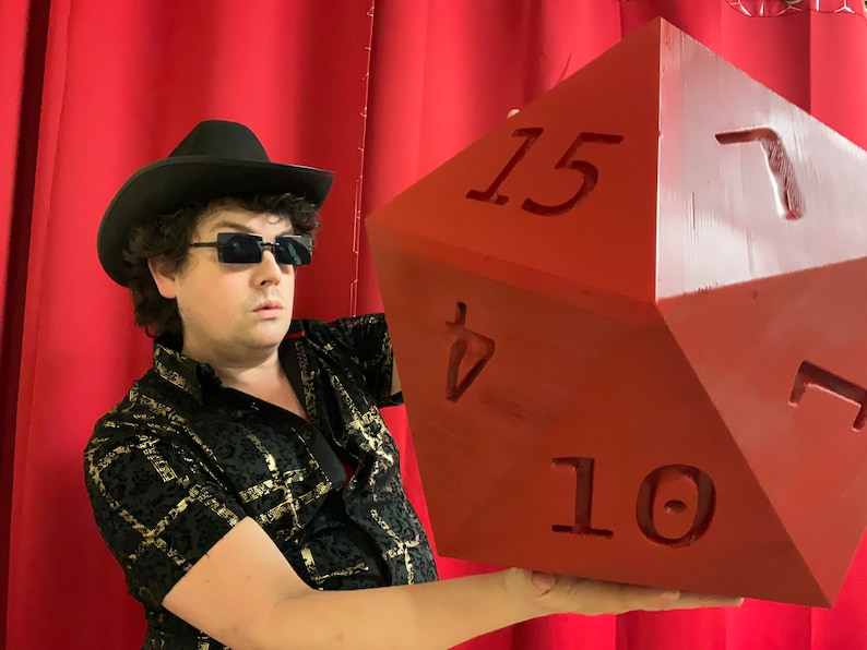 Puede incluir: Una persona con un sombrero negro y gafas de sol sostiene un gran dado rojo de 20 caras con los n&uacute;meros 10, 15 y 7 visibles.