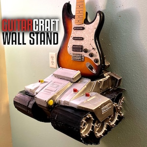 May include: A guitar stand shaped like a tank, painted in a silver and black color scheme. The stand has a yellow and red detail. A guitar is resting on the stand. The text "GUITARCRAFT WALL STAND" is visible in the image.