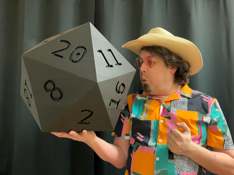 Pu&ograve; includere: Un grande dado a venti facce grigio scuro con numeri neri. Il dado &egrave; tenuto da una persona che indossa una camicia fantasia colorata, un cappello da cowboy e occhiali rosa. Il dado &egrave; un oggetto di novit&agrave; per i giochi da tavolo.