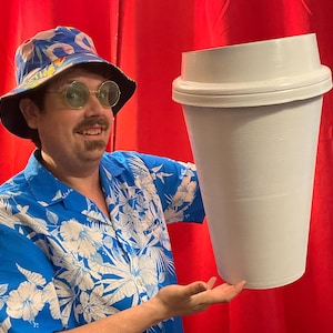 May include: A large, white coffee cup prop with a lid. The cup is held in front of a red backdrop. The person holding the cup is wearing a blue Hawaiian shirt, round glasses, and a bucket hat with a snake pattern.