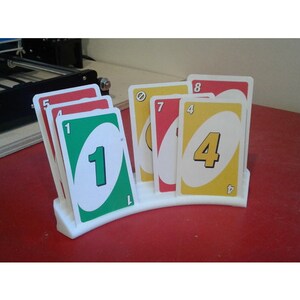 May include: A white plastic card holder with a curved design holds a stack of UNO cards. The cards are red, yellow, green, and blue with numbers and symbols.