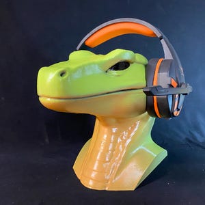 May include: A green and yellow dinosaur head sculpture wearing a black and orange gaming headset.
