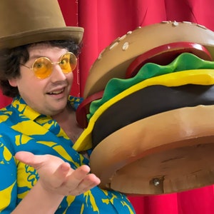 Puede incluir: Un hombre con un sombrero de copa marr&oacute;n, una camisa estampada de color amarillo y azul y gafas redondas sostiene una gran hamburguesa marr&oacute;n y amarilla. La hamburguesa tiene un pan de semillas de s&eacute;samo, lechuga, queso y un tomate rojo.