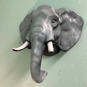 Puede incluir: Una escultura de cabeza de elefante gris con colmillos blancos montada en una pared verde claro.