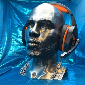 May include: A silver 3D printed bust of a person wearing black and orange headphones. The bust is on a blue background.