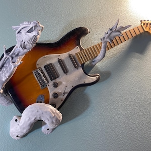 May include: A white, 3D printed dragon sculpture is holding a brown and white electric guitar on a light blue wall.
