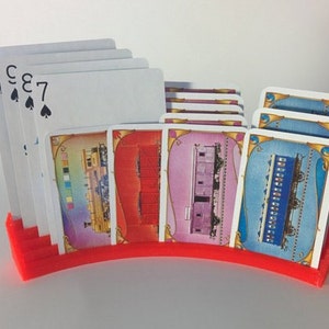 May include: A red plastic card holder with a curved design holds a stack of playing cards and several game cards with train illustrations. The playing cards show the numbers 7, 8, 3, and Ace of Spades.