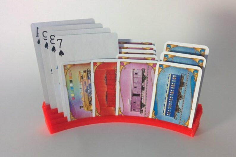 May include: A red plastic card holder with a curved design holds a stack of playing cards and several game cards with train illustrations. The playing cards show the numbers 7, 8, 3, and Ace of Spades.