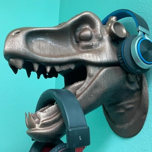 Puede incluir: Una escultura de cabeza de dinosaurio de bronce con un auricular azul y plateado colgando de su boca. La cabeza de dinosaurio está montada en una pared.