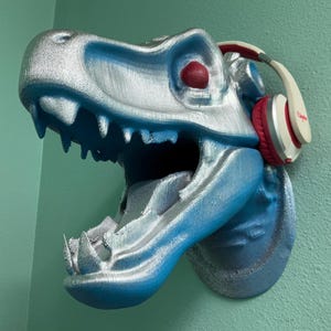 Puede incluir: Una escultura de cabeza de dinosaurio azul y plateada con auriculares rojos. La boca del dinosaurio está abierta y sus ojos son rojos.